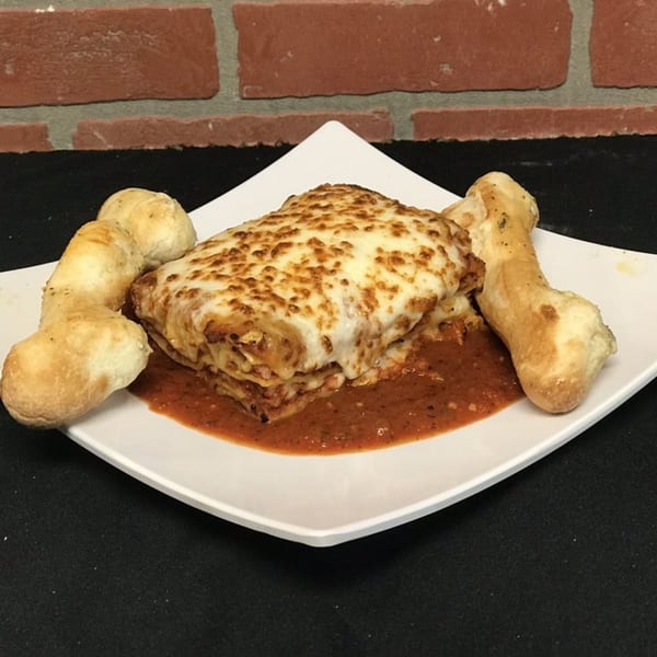 A cheesy lasagna with red sauce and two breadsticks on a white plate against a brick wall.